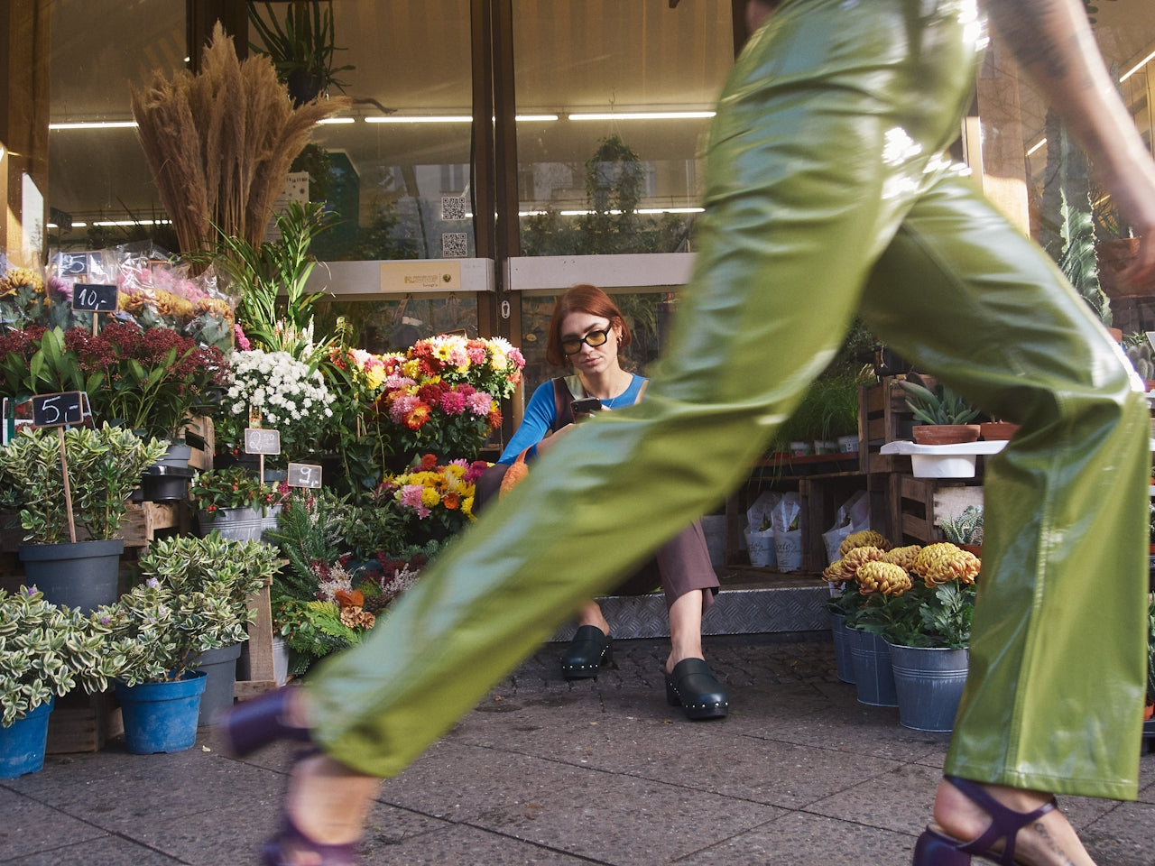 Person in green pants walking past a flower shop with a woman sitting on a bench.